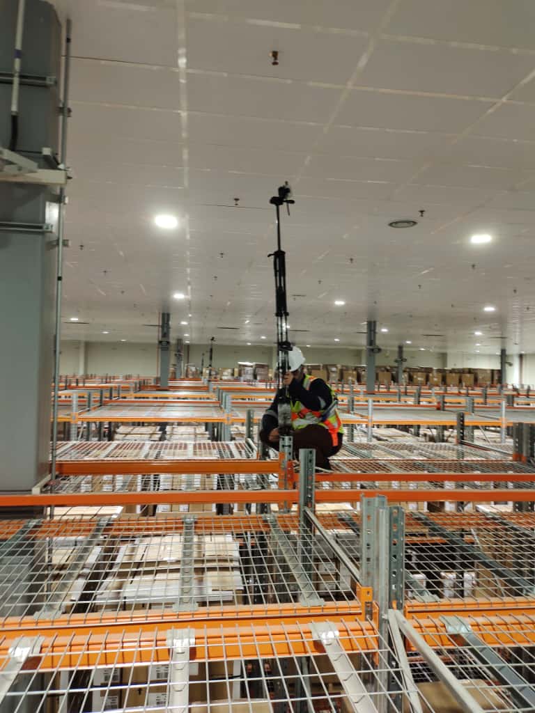 A technician kneeling on warehouse racking beside a tall black sensor mast