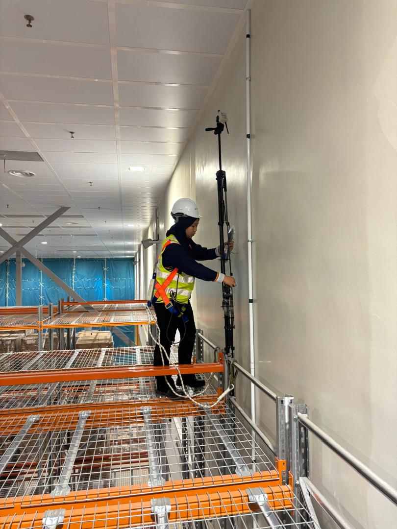 A technician in a hard hat and harness standing on warehouse racking while adjusting a tall instrument mast